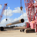 Two large red crawler cranes lifting a heavy steel pipe at an industrial construction site for pile driving. The cranes with lattice booms are positioned on sandy ground under a bright blue sky with clouds. Additional steel pipes and construction equipment are visible in the background, showcasing heavy lifting and infrastructure work.
